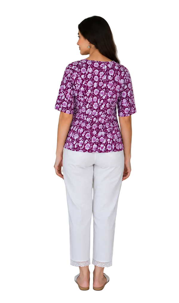 A woman is shown from the back wearing a purple and white floral print short-sleeve top paired with white cropped pants, standing in an indoor setting next to a potted plant and a wooden door.