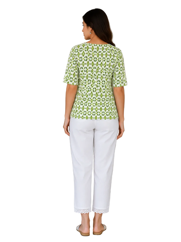 A woman is shown from the back wearing a light green and white patterned short-sleeve top and white cropped pants with lace trim, standing in an indoor setting next to a potted plant and a wooden door.