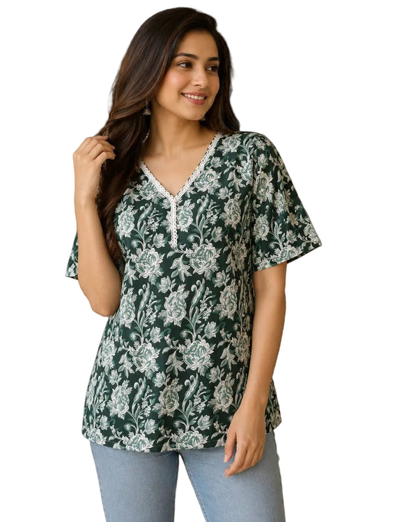 A woman wearing a dark green short-sleeve top with a white floral pattern and lace trim, paired with light blue jeans, standing in a styled indoor setting with plants and home decor.