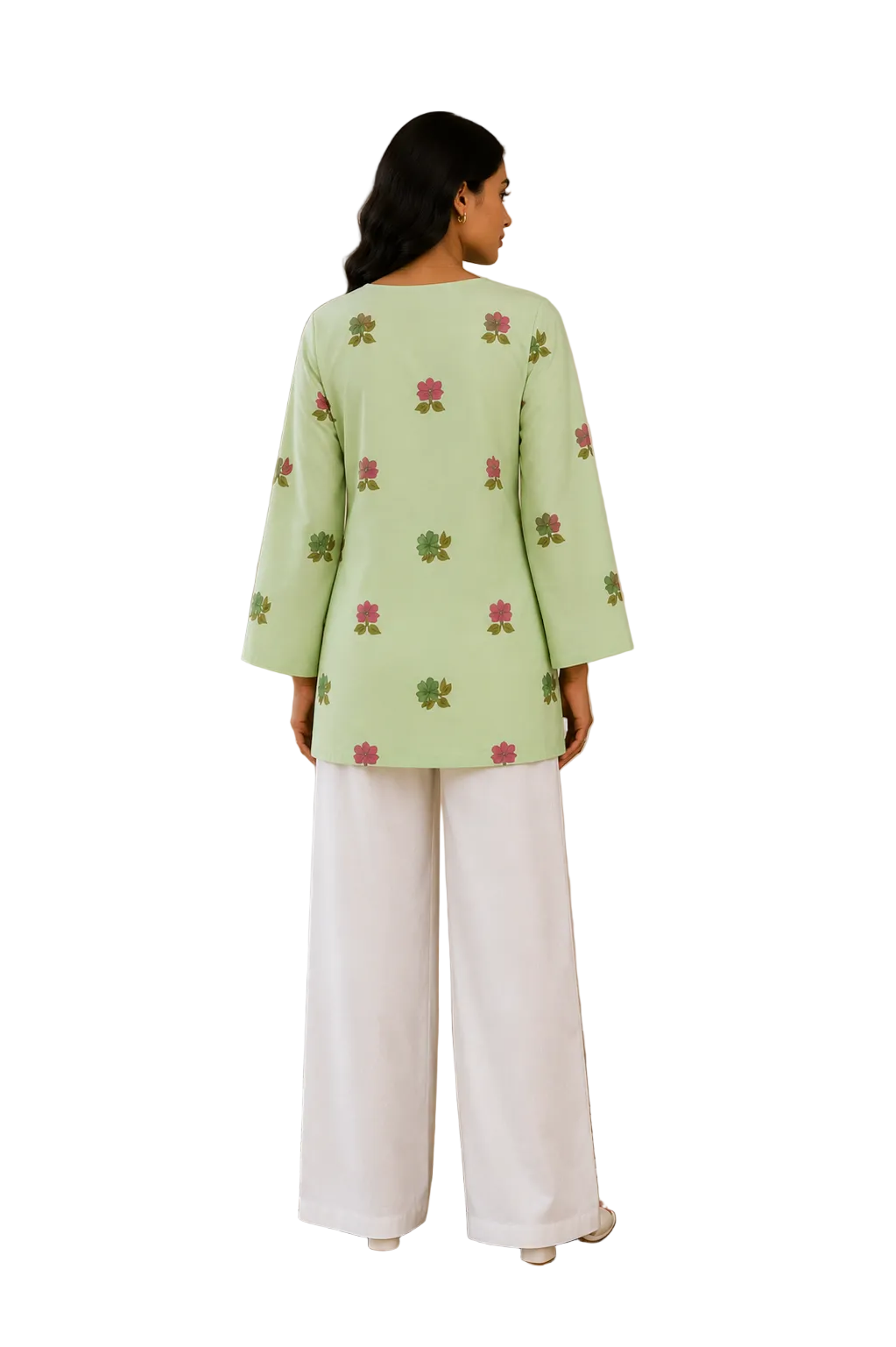 A woman stands with her back to the camera wearing a light green long-sleeve top with pink and green floral embroidery, paired with white wide-leg pants in a warm, neutral-toned room.