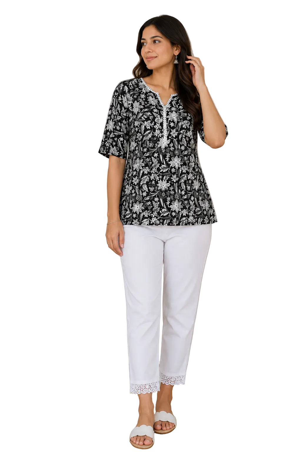 A woman modeling a black and white floral print short-sleeve tunic top paired with white cropped pants, standing in a styled indoor room with a plant, framed wall art, and a wooden side table.