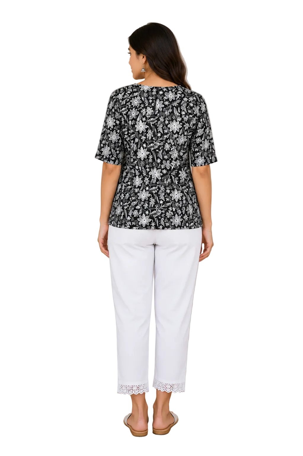A woman viewed from the back wearing a black short-sleeve top with a white floral pattern and white cropped pants, standing indoors on a round woven rug with a plant and wooden table in the background.