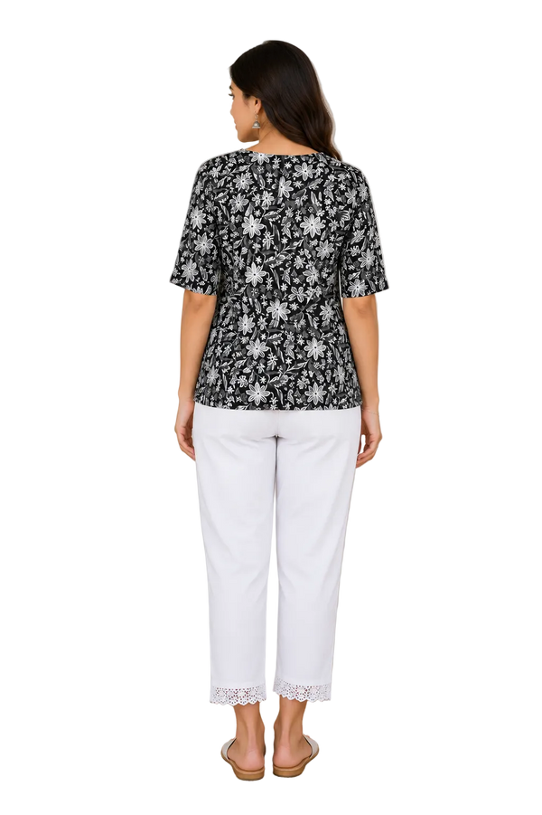 A woman viewed from the back wearing a black short-sleeve top with a white floral pattern and white cropped pants, standing indoors on a round woven rug with a plant and wooden table in the background.