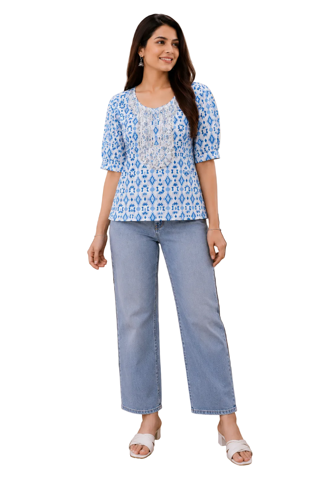 A woman modeling a blue and white patterned short-sleeve top with light-wash blue jeans and white slide sandals, standing in a styled indoor room with a wooden cabinet, plant, and lanterns.