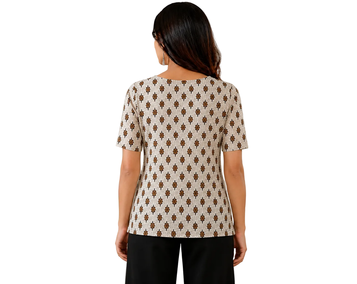 A woman viewed from behind wearing a cream-colored short-sleeve top with a brown geometric pattern, paired with black pants, standing in a brightly lit room with a white cabinet and a vase of flowers in the background.