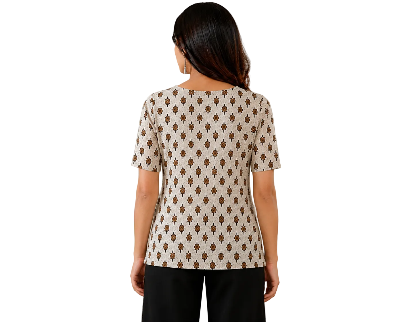 A woman viewed from behind wearing a cream-colored short-sleeve top with a brown geometric pattern, paired with black pants, standing in a brightly lit room with a white cabinet and a vase of flowers in the background.