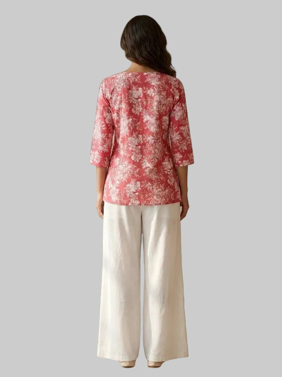 A woman viewed from behind wearing a coral pink and white floral printed top with three-quarter sleeves and white wide-leg pants, standing in a warm beige interior setting.